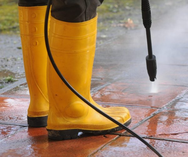 A person wearing yellow rubber boots with high-pressure water nozzle cleaning the dirt in the tiles